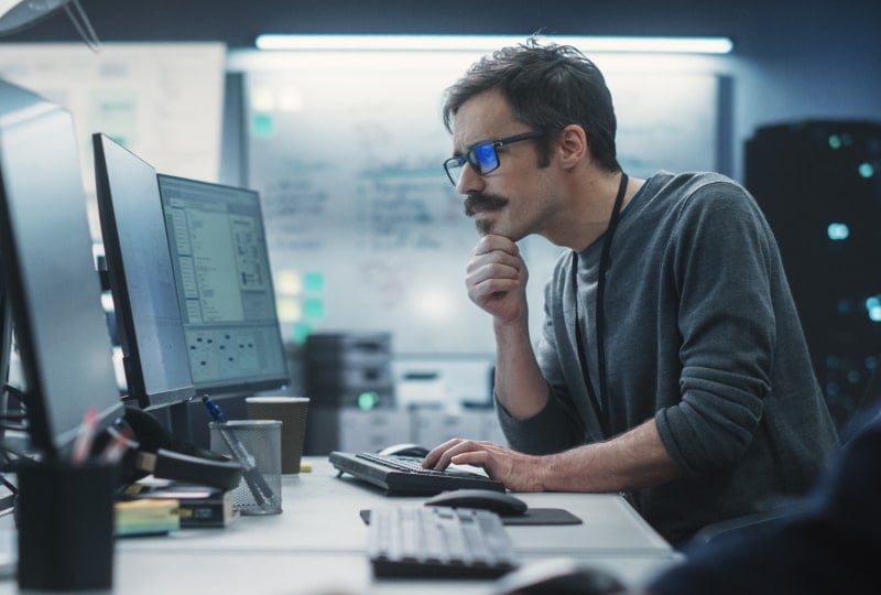 a mustachioed man looking at computer screens