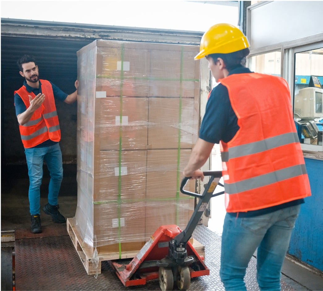 Two workers handling a large pallet wrapped in plastic. One worker is wearing a yellow hard hat and operating a manual pallet jack to move the load, while the other is guiding the process inside a truck. Both are wearing orange safety vests and jeans, indicating a warehouse or shipping environment.