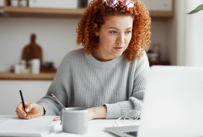 A woman looking towards her laptop while holding a pen over a notepad.