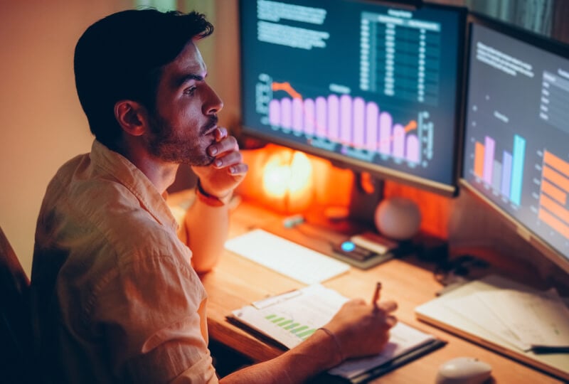 Man taking notes while looking towards a dual monitor setup with charts on each screen.