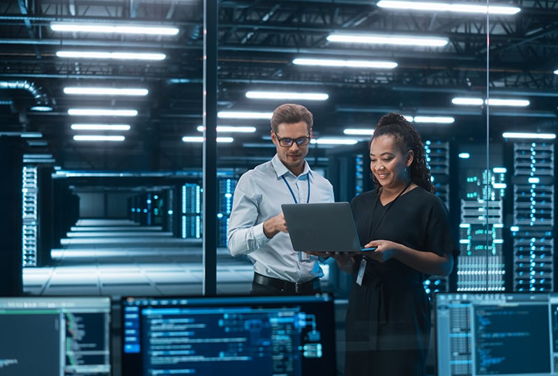 Two people are standing in a modern server room or data center, looking at a laptop together. They are both wearing business attire with ID badges. In the foreground, several computer monitors display code and data, and rows of server racks with blue lighting are visible in the background. The environment is brightly lit with overhead lights, emphasizing a high-tech, professional setting.