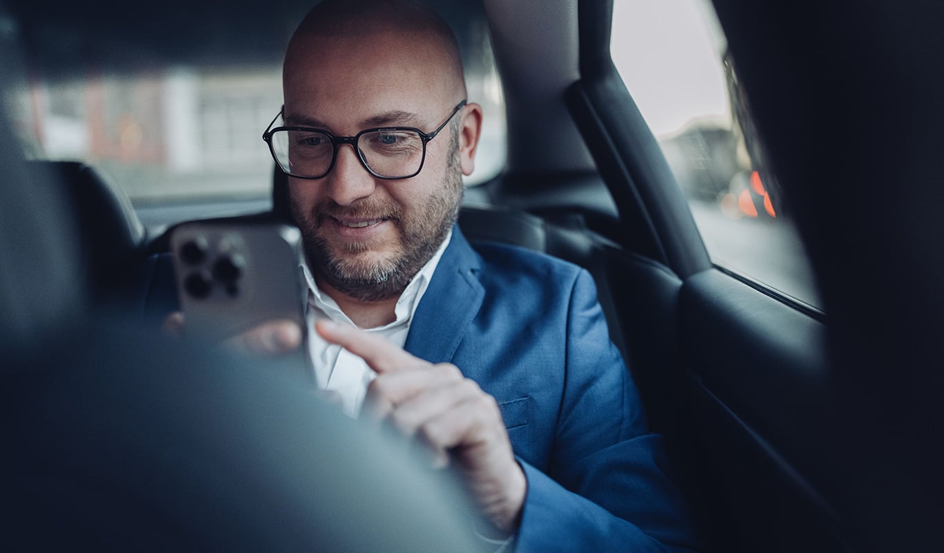 a bald man wearing glasses in the backseat of a car typing on his mobile phone