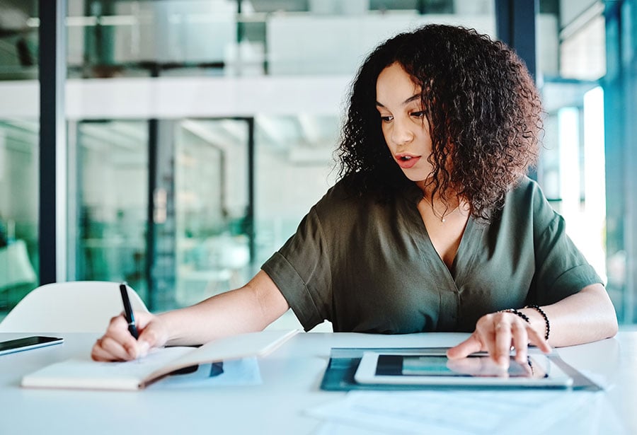 woman writing on notepad while simutaneously scrolling on ipad