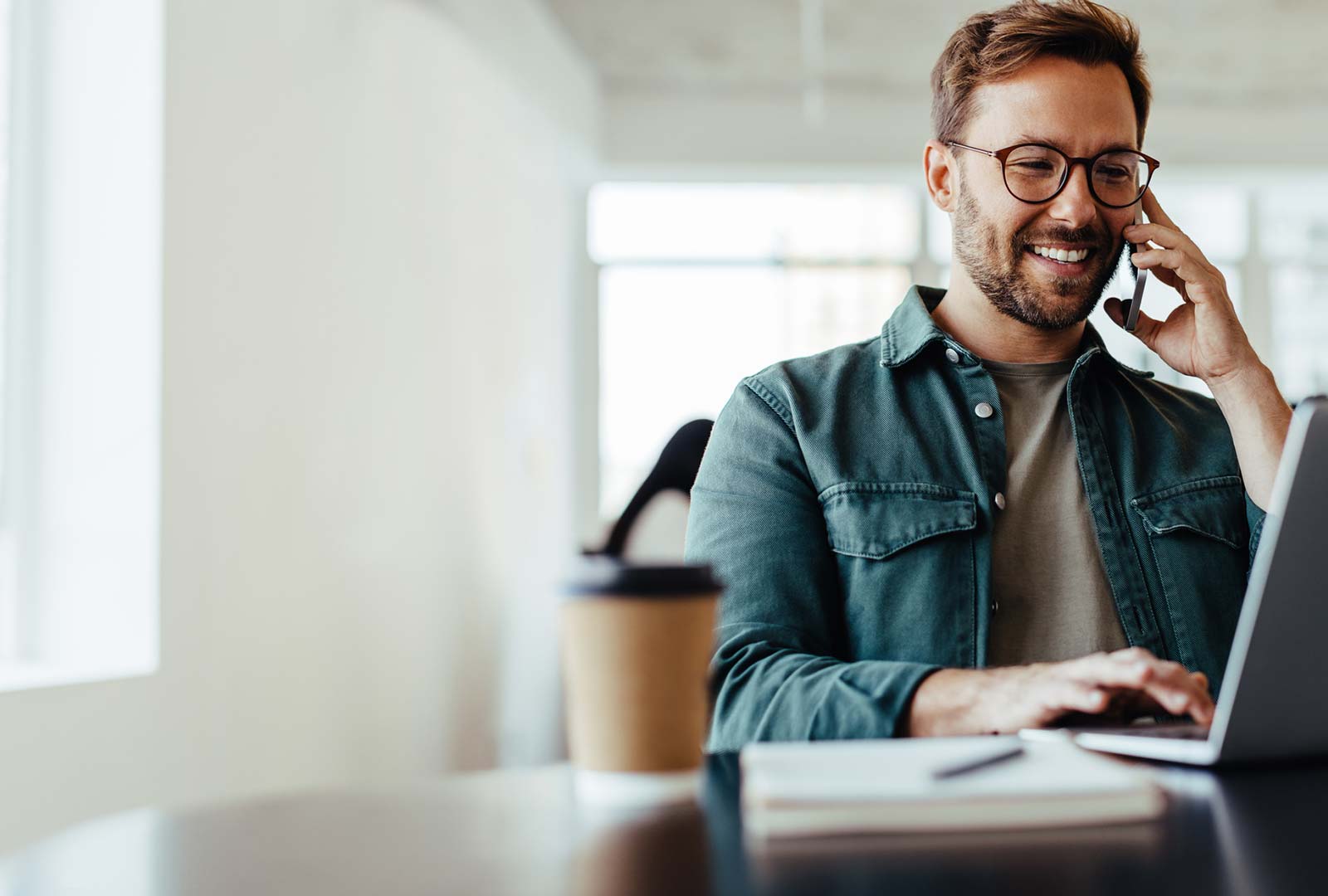 A smiling man holding a phone to his ear while working on a laptop.