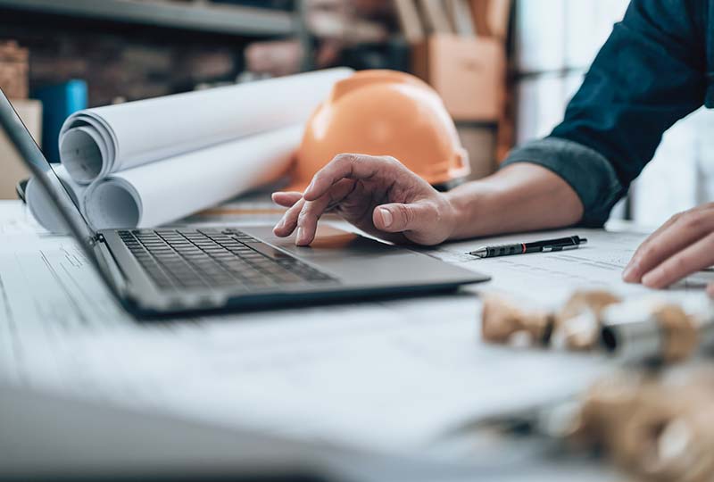 A hand hovering over a laptop trackpad with a hardhat and rolled up documents in the background.