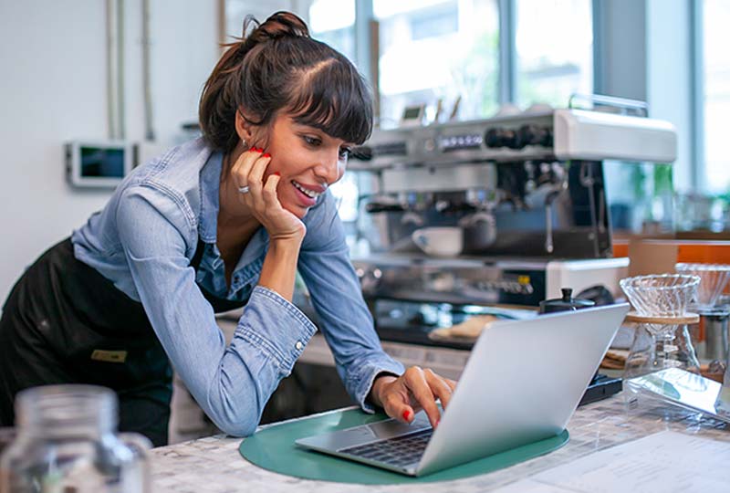 A smiling woman leaning and resting her arm on a countertop in a cafe while using a Chromebook.