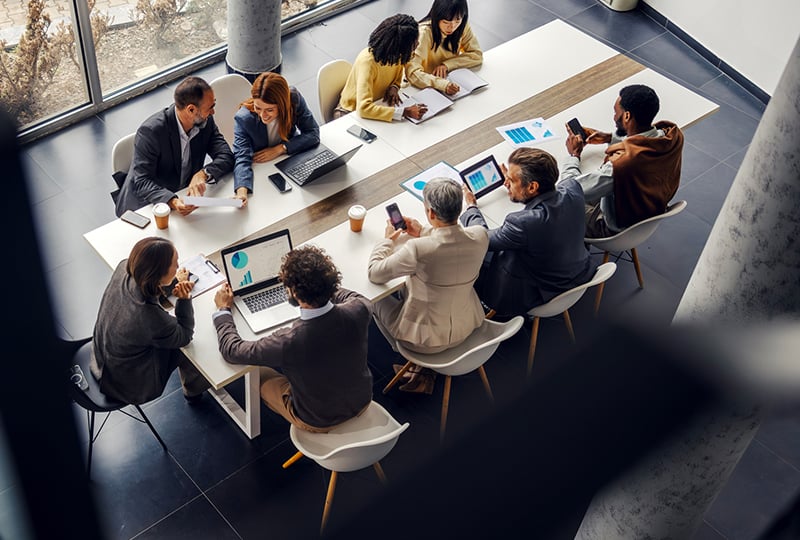 Overhead view of a large conference table with a group of people seated around it, working on laptops, reviewing printed charts, and using mobile devices during a collaborative meeting.