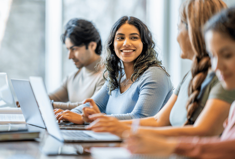 Smiling female university student interacting with a laptop in a classroom with other students.