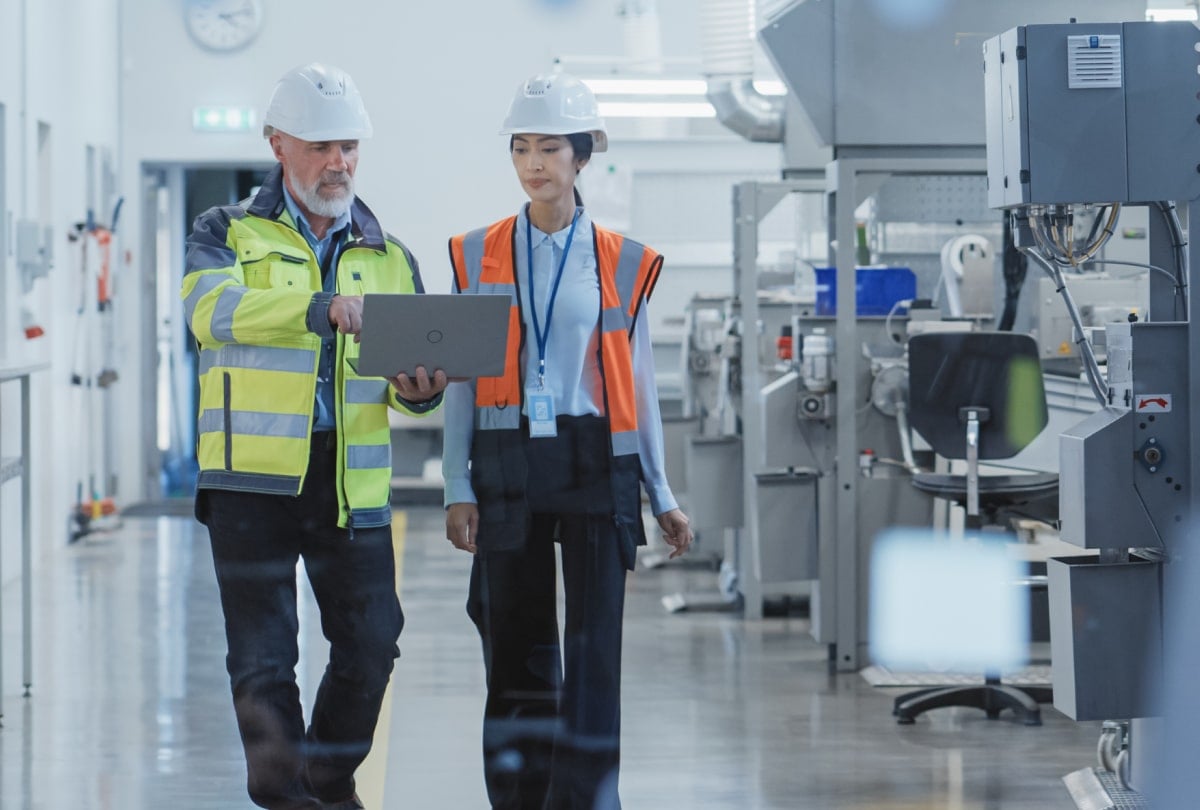 一名女性和男性穿着警示背心,走在生产线通道里,讨论笔记本电脑上显示的数据。