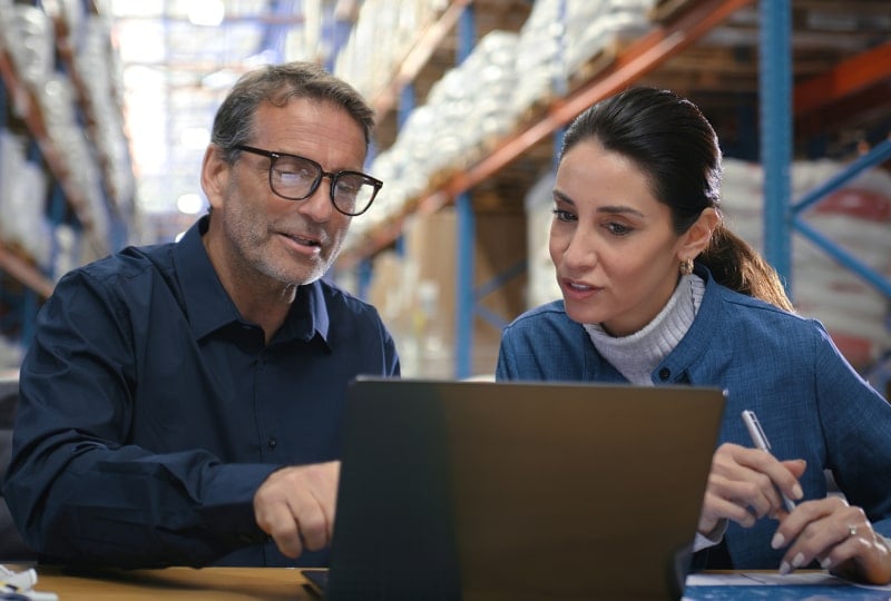 一男一女在仓库内看着笔记本电脑上的显示内容。