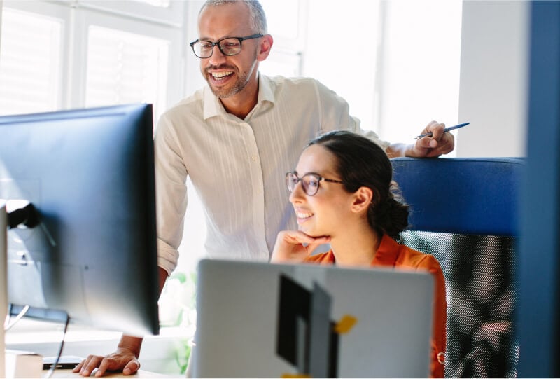 A man and woman smiling while looking at a computer monitor on the woman's desk.