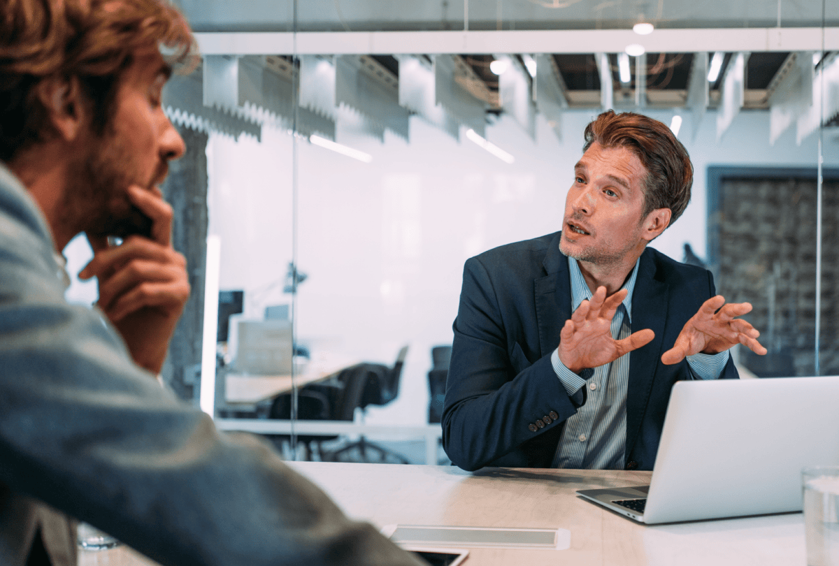 Two men in a meeting room where one of them is gesturing towards a laptop.