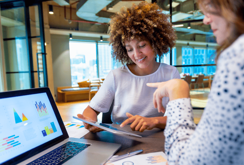 Two women sitting and having a meeting, while interacting with a tablet computer held in one of the woman's hands.