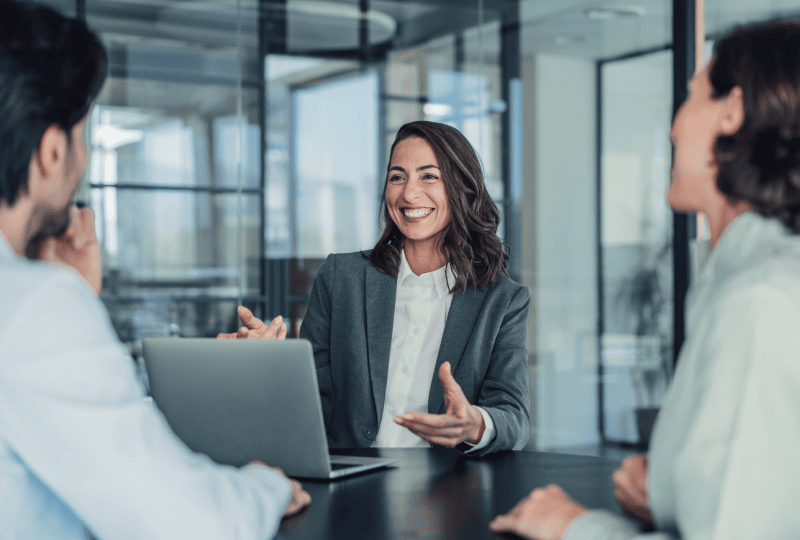 A woman smiling and gesturing towards a man and woman sat in front of her in a meeting.