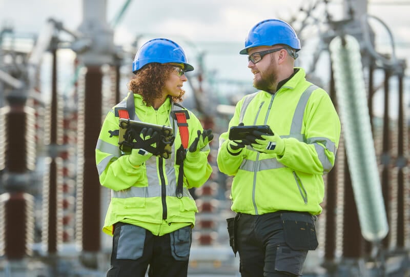 A man and woman both wearing high visibility jackets and hardhats talking and interacting with tablet computers near electric superconductors.