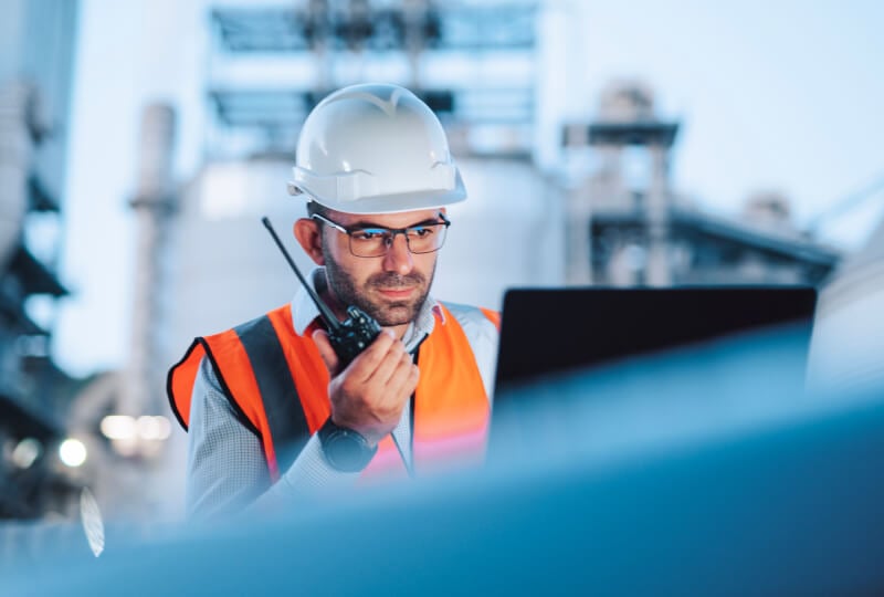 Man in a high visibility jacket and hardhat talking on a walkie-talkie and interacting with a computer monitor.