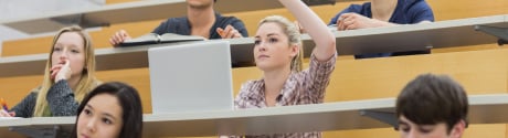 Female student working on a laptop and raising her hand in a lecture hall