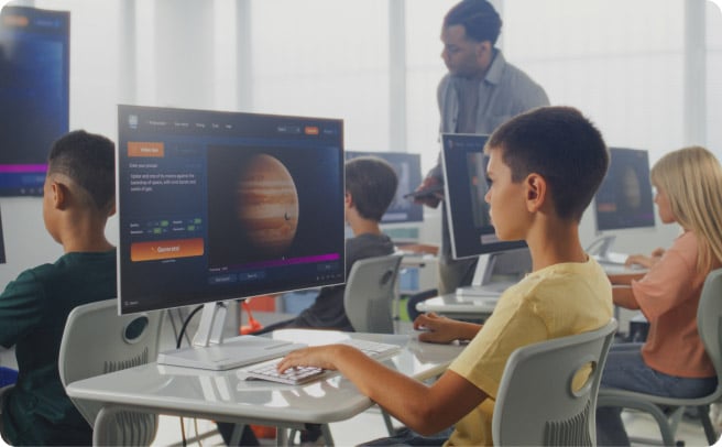 A male student sitting at a computer in a classroom looking at a screen, while a teacher stands in the background.
