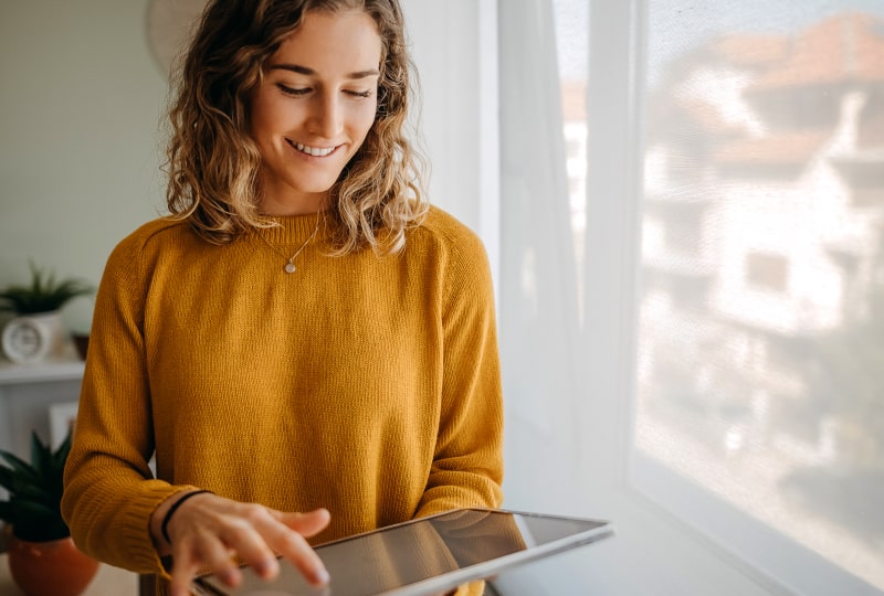 Smiling woman in a yellow sweater interacting with a tablet held in her left hand.