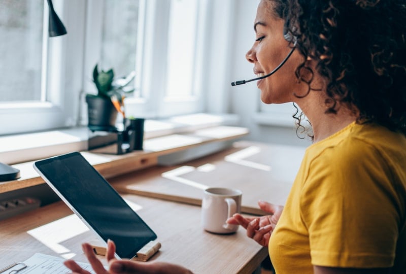 Side shot of a smiling woman wearing a chat headset gesturing towards a tablet device.