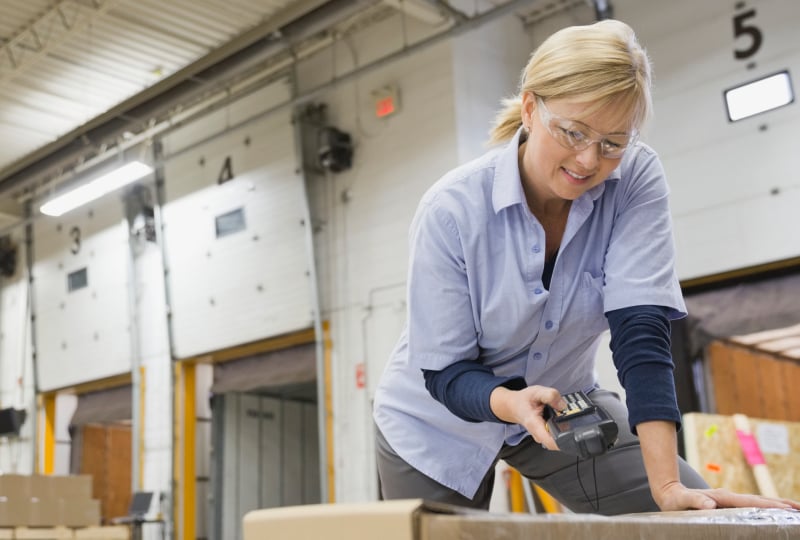 Female warehouse worker leaning over and scanning a box with a mobile scanning device.