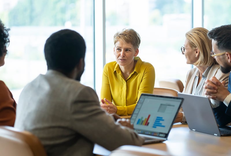 Team meeting around a conference table with four people, one person speaking while others listen, laptops open displaying charts and data.