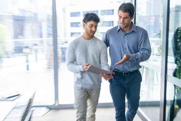 Two men walk through a bright office hallway while looking at a tablet together