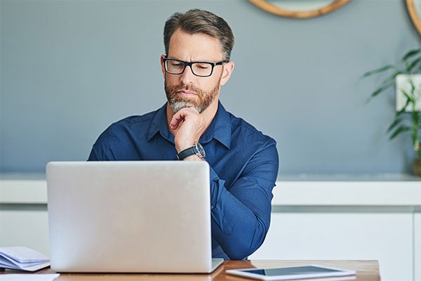 Bearded man wearing glasses sits in front of a laptop with his chin resting on one hand