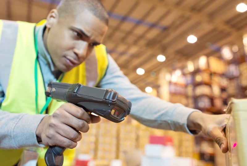 Man in a warehouse wearing a yellow reflective vest scanning a box with a mobile scanning device.