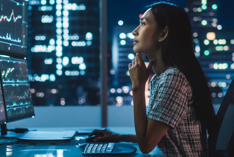 A woman sitting in an office at night looking towards a dual monitor display on her desk.