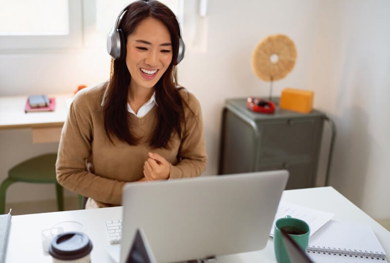 A smiling woman wearing headphones and interacting with a laptop in front of her.