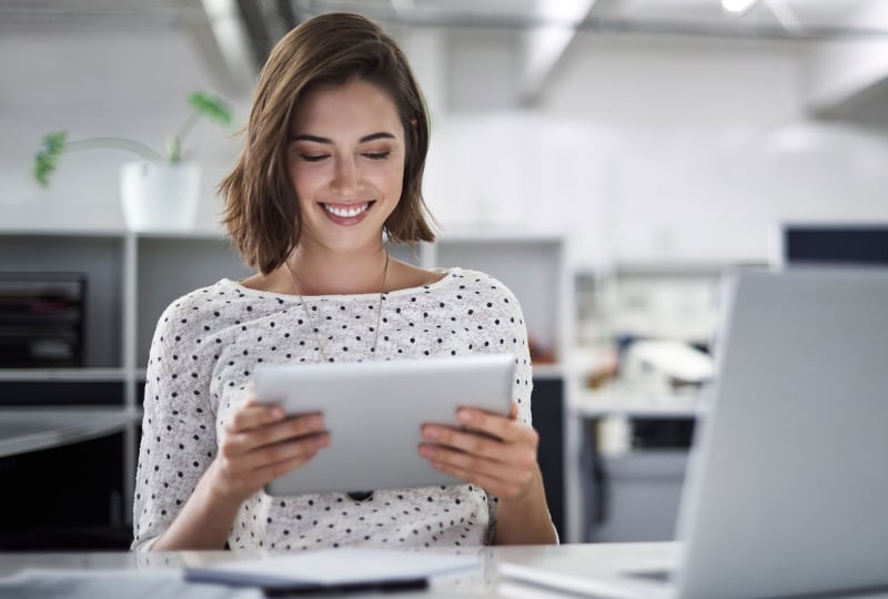 Smiling woman sitting at a desk interacting with a tablet computer in her hands.