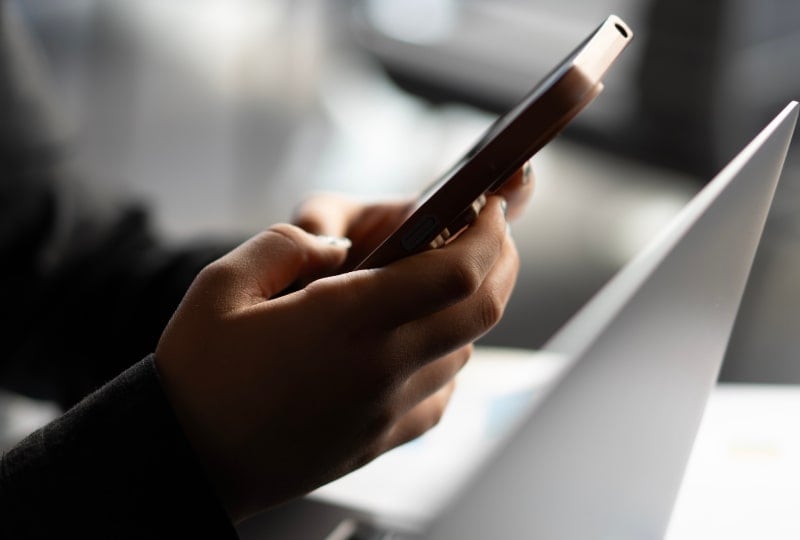 closeup of a person using a mobile phone next to a laptop