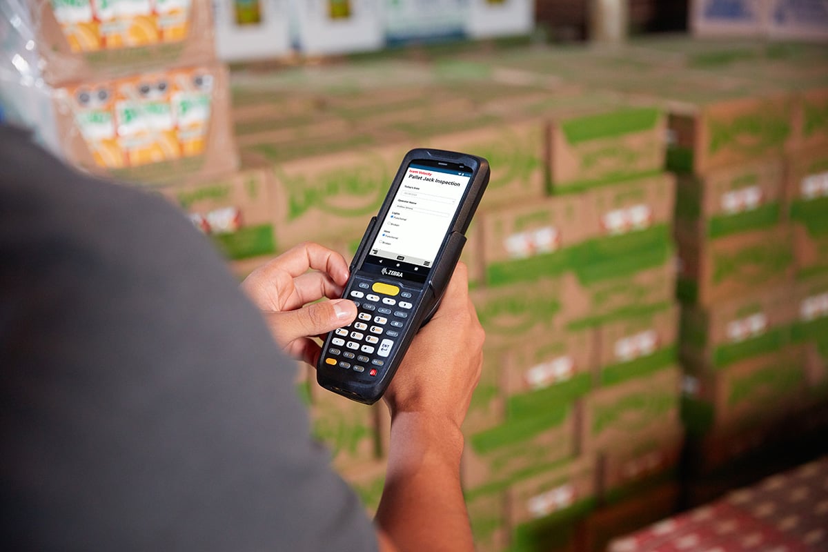 A person is holding and using a handheld barcode scanner or mobile computer device labeled "Zebra" in a warehouse or storage area. The screen displays a form titled "Pallet Jack Inspection." In the background, there are stacks of cardboard boxes. The image suggests inventory management or equipment inspection in a warehouse setting.