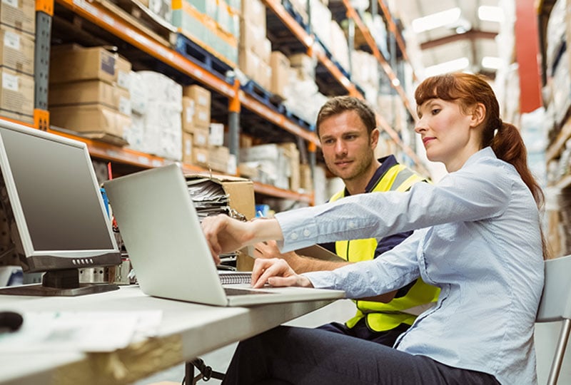 Two people sitting at a desk in a warehouse. One person is wearing a safety vest while the other is pointing at a laptop screen. There are shelves filled with boxes and inventory in the background, and a computer monitor is on the desk.