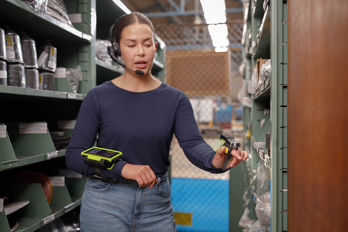 A woman working in a warehouse, wearing a headset, a wearable barcode scanner on one hand, and a mobile device strapped to their arm. They are standing between shelves filled with various items and appear to be scanning inventory.