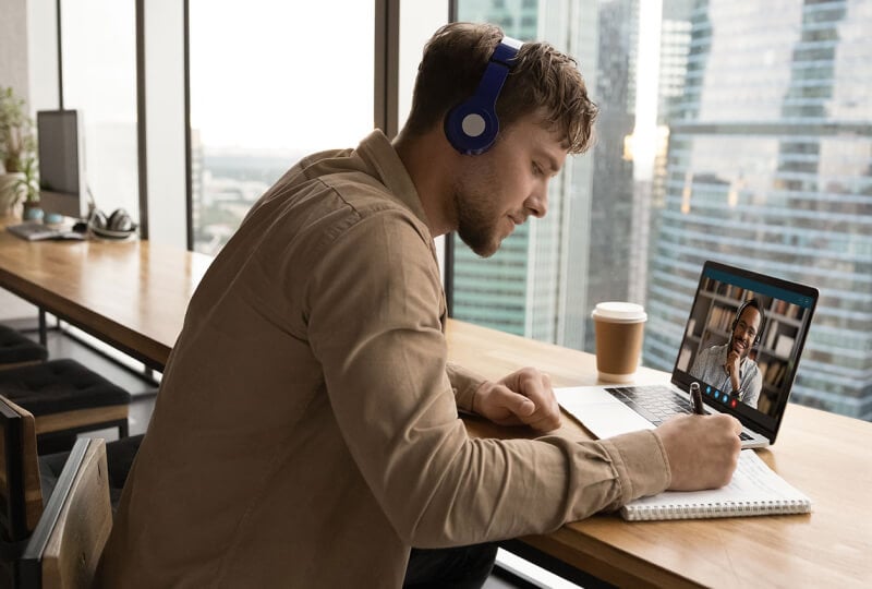 A man in the upper floor of a skyscraper building seated near a window taking notes while listening in to a video meeting on his laptop.