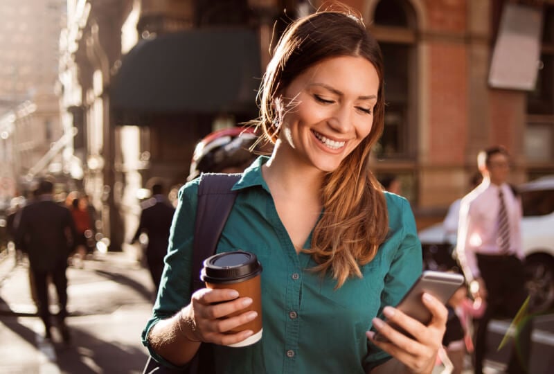 Smiling woman on a busy city street holding a coffee in one hand and her phone in another.